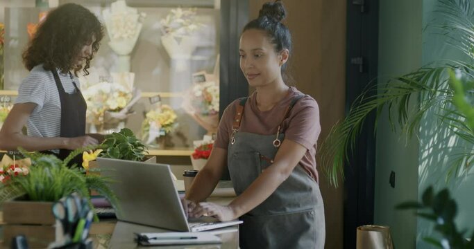 Young woman flower business owner using laptop while female employee bringing boxes with plants in shop. Retail and commerce concept.