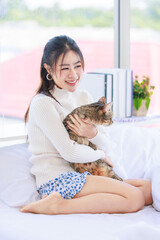 Asian pretty female teenager girl in turtleneck sweater wearing headphones siting smiling on white clean sheet bed and holding tabby short hair cat