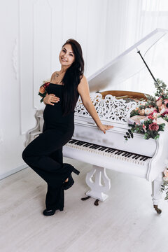Vertical image pregnant woman strokes hugging the belly tummy abdomen enjoying pregnancy standing near grand piano dressed black suit. Future family, baby infant expecting child inside

