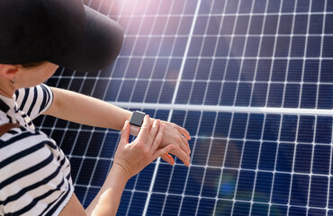 rear view female person wearing cap standing in front of big solar panel and remotely control it with her digital watch on hand.