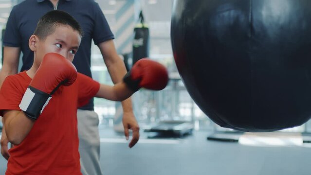 Small asian boy in boxing gloves trains to punch bag in gym with trainer, side view. Father watches his son's boxing training at gym. Sports concept