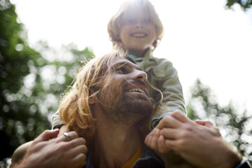 Closeup portrait of happy young father carrying son on shoulders outdoors in sunlight