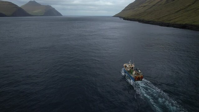 Aerial view at close range and with tracking from the back of a fishing boat that sails through a fjord in the Faroe Islands and where the great mountains can be seen.