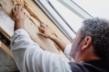 Carpenter seen over shoulder fixing insulation material on roof window