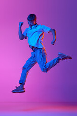Fototapeta na wymiar Full-length image of young guy in casual clothes and headphones jumping over gradient pink purple background in neon light. Concept of human emotions, youth, lifestyle, fashion, facial expressions, ad