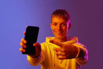 Smiling young man pointing on mobile phone screen against gradient purple background in neon light. Concept of human emotions, youth, lifestyle, online communication, communication, social media, ad