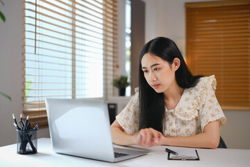 Fototapeta na wymiar Focused young Asian woman looking at laptop screen, working or studying online at home
