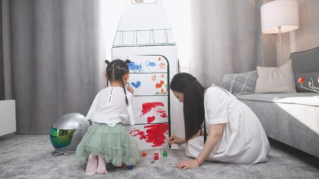 Asian woman with her daughter play in the living room at home, sitting on the floor, girl together with mother paint on a cardboard model of a spaceship with paints.