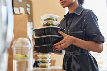 Close up of young African American woman unpacking food delivery order at home and holding stack of containers - obrazy, fototapety, plakaty