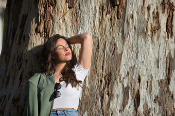 Young and beautiful woman, Hispanic, brown hair, with green jacket and jeans, eyes closed, alone, relaxed and calm, leaning against the trunk of a huge tree. Concept of solitude, relax, calm.