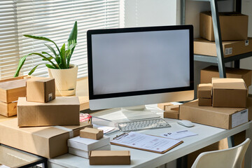 Desktop computer with blank screen standing among packed boxes containing orders of online clients on workplace of warehouse manager