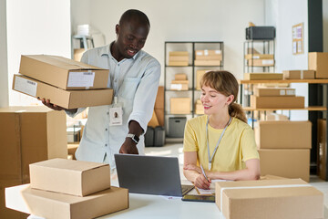 Young African American male manager of warehouse with stack of boxes pointing at laptop screen while showing his fremale colleague
