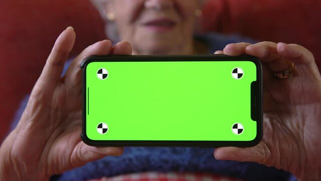 Old woman at home using a mobile phone with green screen. Mockup device to add elements. Wrinkled hands of grandmother holds a smartphone. Concept of technology