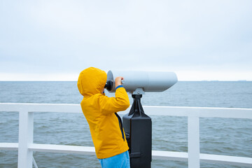 A child in a yellow jacket is standing on the pier, looking through binoculars at the sea. Cold...