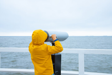 A child in a jacket stands on a white pier. looking through binoculars at the sea. Cold weather....