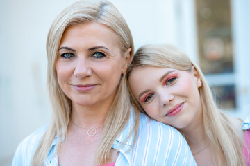 Beautiful family portrait of happy blonde mom and daughter smiling at camera, standing on street against white wall