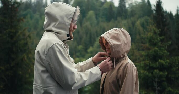 The guy fastens the girl's jacket and kisses her during the rain in a mountain coniferous forest. Happy couple against green forest background