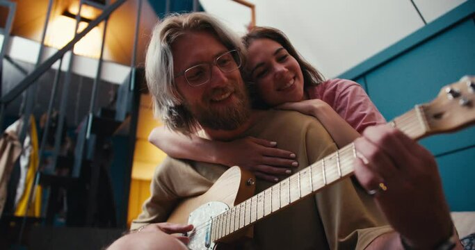A guy a girl spends time together a blond guy with a beard in glasses plays the electric guitar a brunette girl hugged him and listens