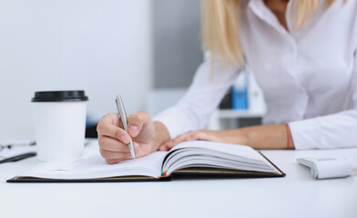 Female hand holding silver pen ready to make note in opened notebook sheet. Businesswoman coffee cup workspace make thoughts records at personal organizer white letter collar conference signature