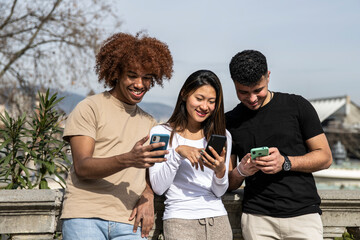 Three young multiracial people texting with smartphones standing in a park. Diverse group smiling and using phones outdoors on a stone balcony