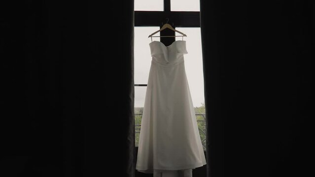 A beautiful white dress hangs in front of the window behind the curtains. Interesting composition