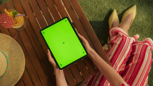 Top View of Anonymous Female Using a Digital Tablet Computer with Mock Up Green Screen Chromakey Display in a Garden. Woman Watching a Recorded Conference While Relaxing and Having a Cocktail Outdoors
