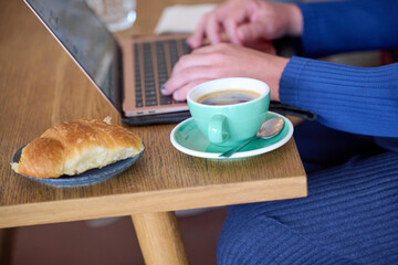 Person working on laptop with croissant and cup of coffee on a wooden table in a cafe