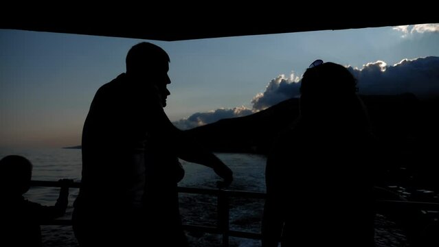 The silhouette of a young couple embracing on a ship in the evening, they are enjoying a tour of the coast with mountains. Cruise on the sea in summer.