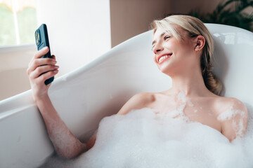 Naklejka premium Young beautiful sexy woman having fun while lying in bathtub full of foam at home. Charming smiling model relaxing in luxury bath interior. Female holding smartphone, taking selfie photos, video call