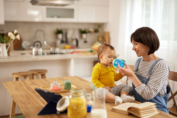 Mother giving her baby boy to drink water from baby bottle at home