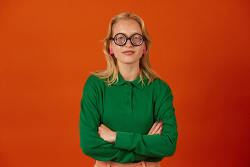 Young woman smiling with folded hands, wearing glasses, green shirt, isolated on orange color background