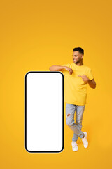 Best deal promotion. Smiling indian teenage guy leaning on huge smartphone with blank screen, pointing finger at empty display and smiling, arab man advertising new app or website, mockup