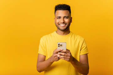 Portrait of cheerful young Indian guy sending message on mobile phone over yellow background. Happy Arab man using smartphone, enjoying chatting online, messaging in social networks