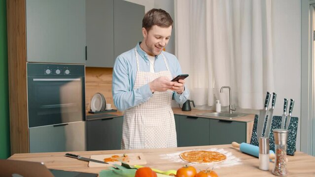 joyful man wearing apron prepares pizza in modern kitchen. Male puts tomatoes on the dough with the sauce and takes pictures with his phone