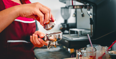 Hand of a barista holding a portafilter and a coffee tamper making an espresso coffee. Barista presses ground coffee using a tamper in a coffee shop
