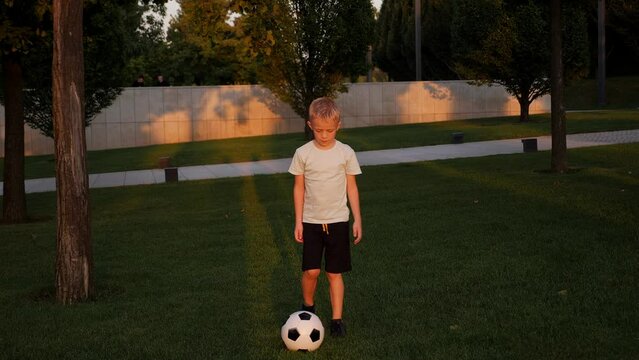 An eight-year-old boy in sports clothes is engaged with a soccer ball in a city park in the summer at sunset. The child is engaged in football.