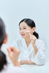 아시아 한국의 검은머리의 젊은 여자 모델이 밝은 표정으로 거울잎에 앉아 샤워가운을 입고 거울을 보며 핑크색 마사지 롤러로 얼굴 마사지를 하고있다.