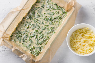 Spinach sorrel pie dough in a baking dish with parchment, a bowl of grated cheese on a light blue background, top view. Cooking step, step by step