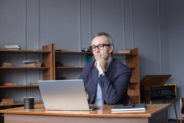 Portrait of mature grey haired businessman in glasses and suit sitting by the wooden table and looking on his laptop