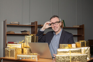 Fototapeta na wymiar Portrait of mature excited businessman in glasses and blue suit sitting by the table with laptop and a lot of gifts around