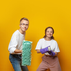 Cleaning concept. Happy young couple man and woman with household tools in hands posing against yellow studio wall banner background