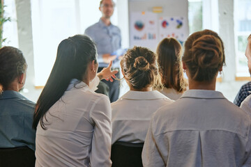 Rear view of woman pointing something in front of her while listening presentation against defocused leader, who teaching team of business people.