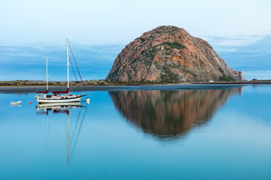 Morning view of Morro Rock in Morro Bay, California. Harbor in foreground with sailboat. Mirror reflection on the water. 

