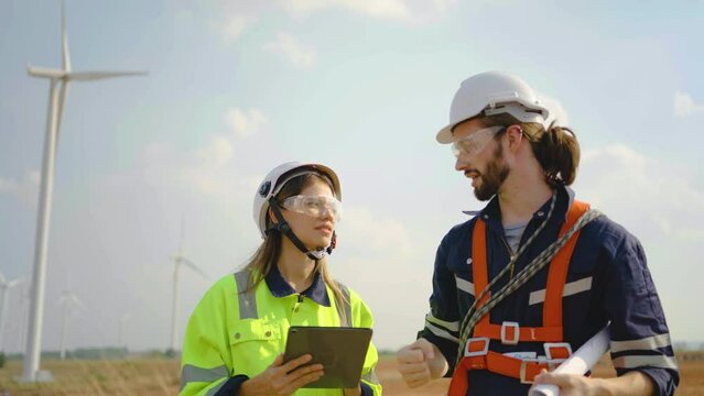 Front view of two engineers caucasian man and woman in uniform and goggles talk about the blueprint and use tablet working at wind turbine farm, wind turbine generate electricity, Renewable energy