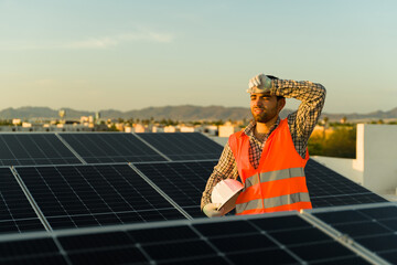 Young man installing solar panels on top of a roof and looking very tired while wiping sweat from his forehead