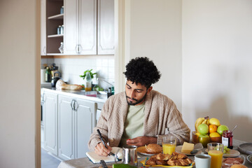 Young man eating breakfast and writing in planner