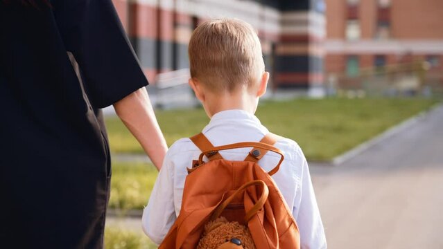 Back view mother leads her son by hand to school. Family is holding hands. Baby boy in school uniform. White shirt and backpack. Preschool child goes to first grade. September. New academic year.