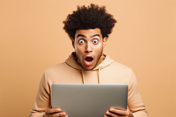 Surprised African teen guy holding laptop pointing at computer advertising wow offer. Amazed ethnic teenager student boy using pc feeling shocked isolated on beige background