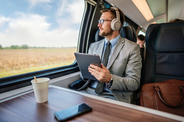 Handsome businessman is having a good time while traveling by high-speed train. He is using laptop computer and wireless headphones for online communication, gaming and entertainment. - obrazy, fototapety, plakaty