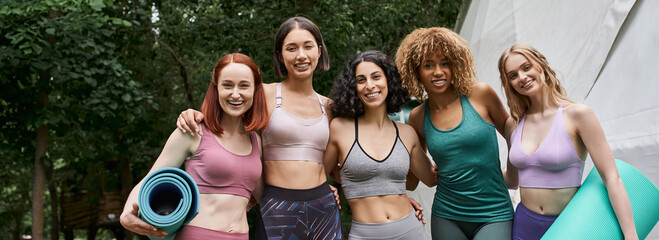 women retreat concept, pleased multiethnic girlfriends with yoga mats looking at camera in park